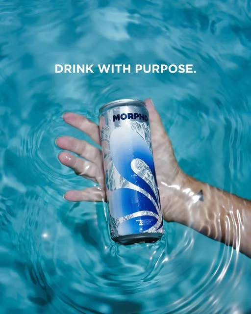 A hand holding a MORPHO can in clear blue water, with the words Drink with purpose